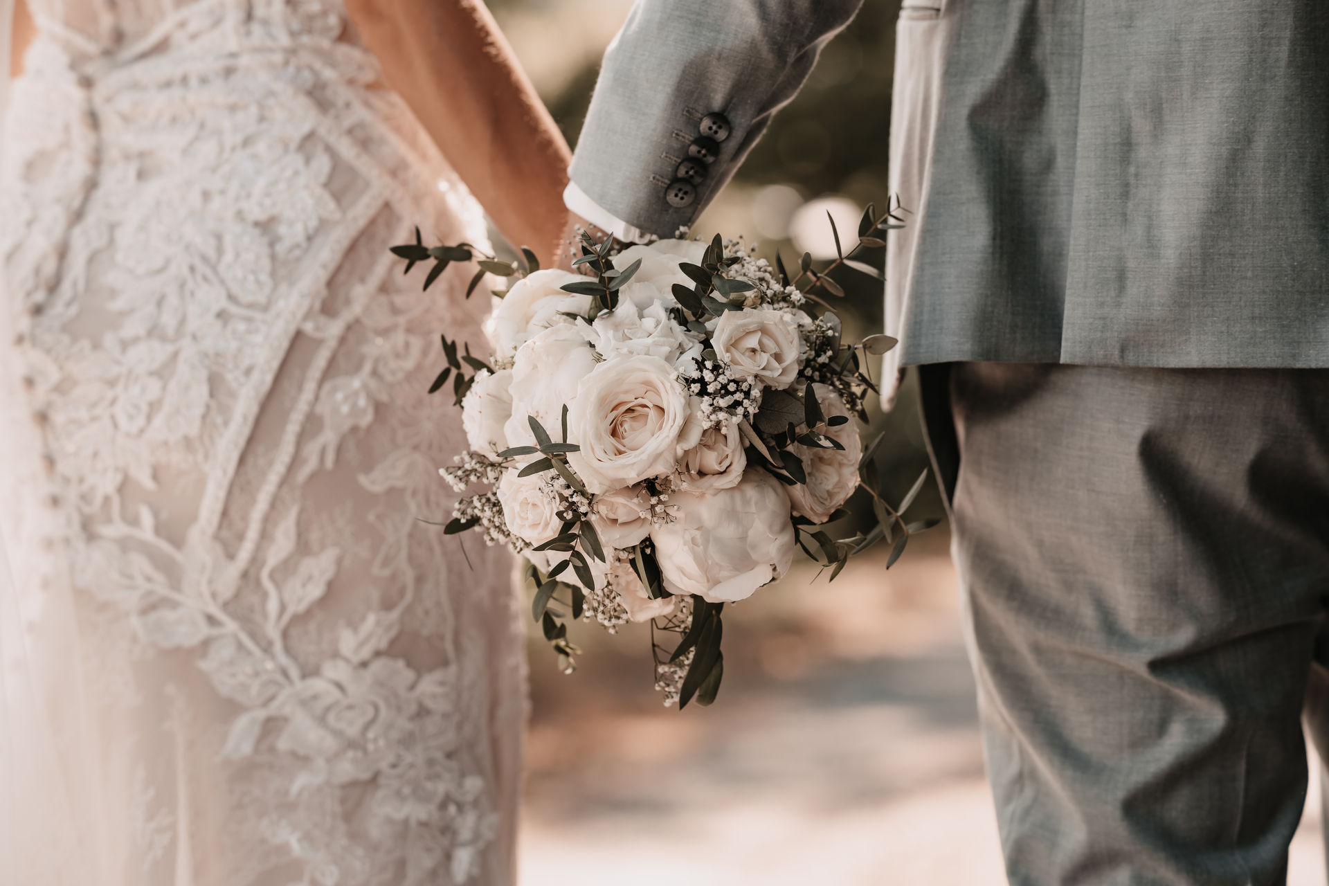 2 - 20230623-DSC02718 Nahaufnahme eines Brautpaares von hinten – die Braut im detailreichen Spitzenkleid hält einen eleganten Hochzeitsstrauß mit weißen Rosen, während sie die Hand ihres Partners in grauem Anzug hält – romantische Hochzeitsfotografie in Bühl.