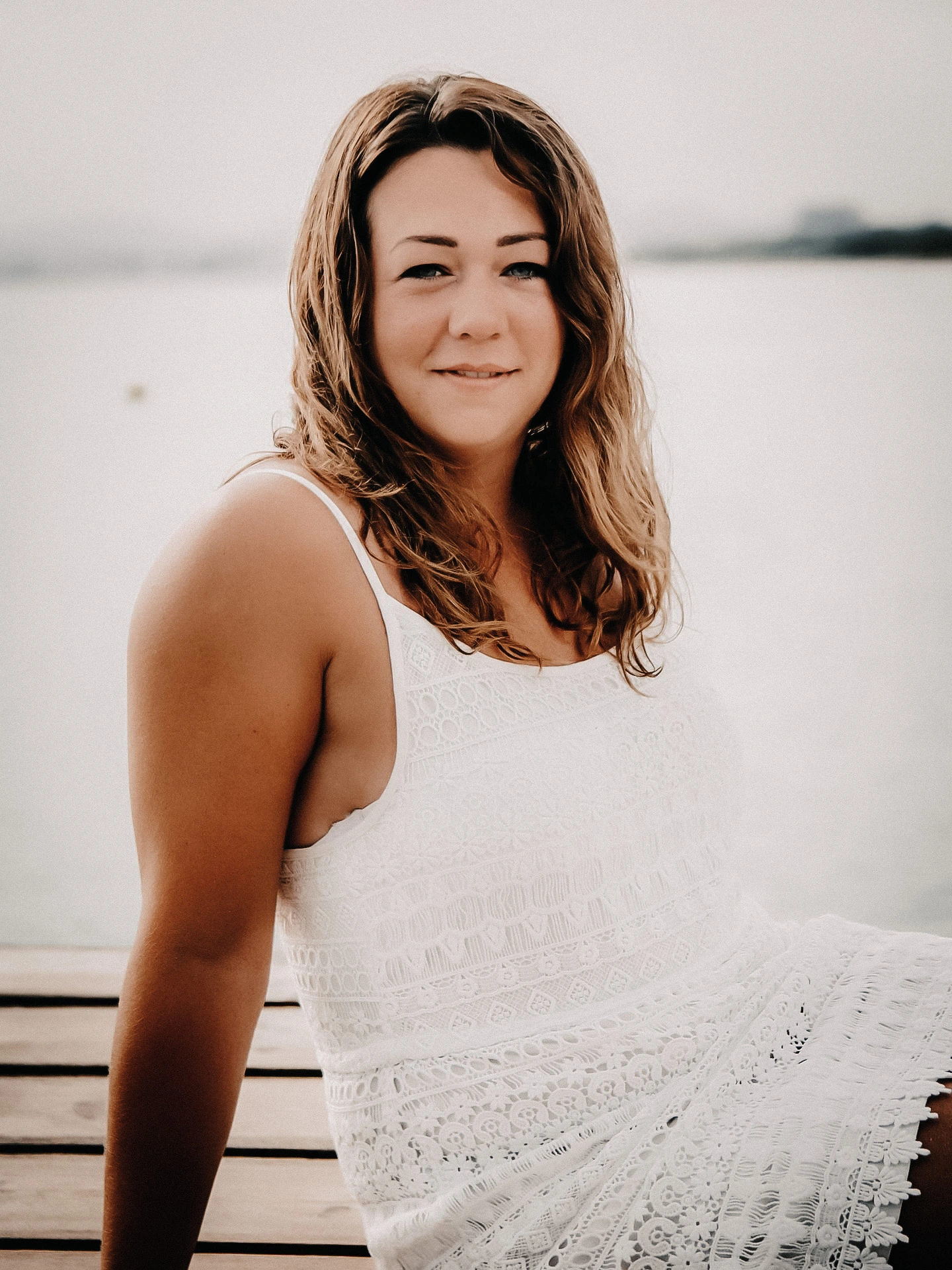 Belio-Photography-Stefanie-Miller Steffi Miller mit welligen, braunen Haaren sitzt in einem weißen Spitzenkleid entspannt auf einem Holzsteg am Wasser und lächelt ruhig in die Kamera – Stefanie Miller, Fotografin bei Belio Photography in Bühl.